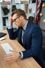 tired young businessman in eyeglasses gesturing by hand at table with computer keyboard and mouse in office