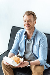smiling young businessman having lunch with burger while sitting on sofa in office