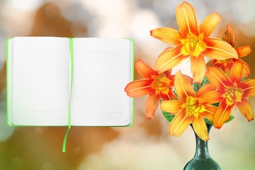 Beautiful live lily bouquet bouquet in ceramic vase with opened note book with blank place for your information on left on nature leaves and branches bokeh background.