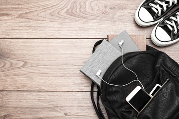 Black leather backpack with books, phone, player with headphones and sneakers on a wooden background.