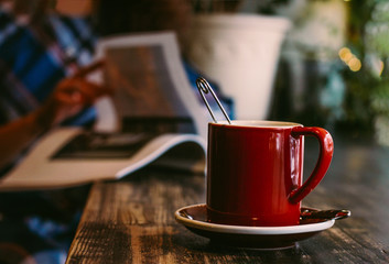 Atmospheric photo with red mug on wooden table in cafe on the background of girl with magazine, cozy winter day