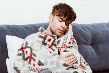 sick man wrapped in blanket sitting with cup of tea in bed at home