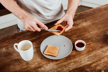 partial view of man spreading toast by jam at table with coffee cup in kitchen at home
