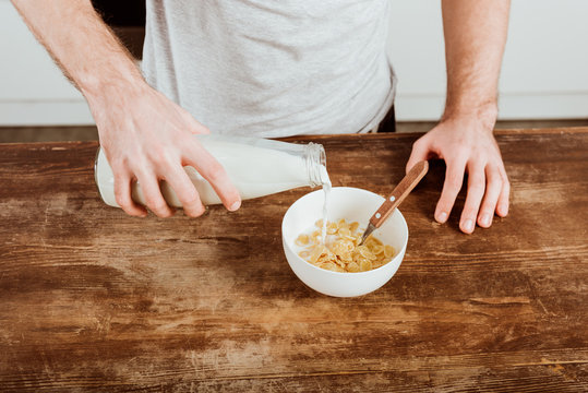 cropped image of man pouring milk into bowl with corn flakes at table with laptop in kitchen at home