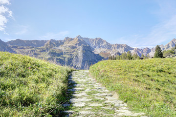 Fototapeta na wymiar A paved path with cloudy sky, high mountains, fir and pine tree forests and green pastures in Val d'Otro, Piedmont region, Alps mountains, Italy