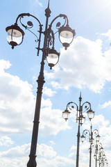 Row of graceful triple street lamp posts under sunlight against blue sky with clouds