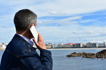 Man talking on the phone. White smartphone, blue clothes. Promenade with rocks and beach. Cloudy sky, La Coruña, Spain.