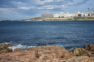 Fototapeta na wymiar Angler with a white fishing rod in the rocks. Blue water with foam, Torre de Hercules and city. La Coruña, Galicia, Spain.