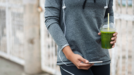 Detail of woman holding green detox smoothie and smartphone while taking a workout rest. Urban...