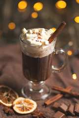 Hot chocolate in a transparent mug with whipped cream and cinnamon sticks, spices, nuts and cocoa powder on a rustic wooden background, selective focus, vertical composition