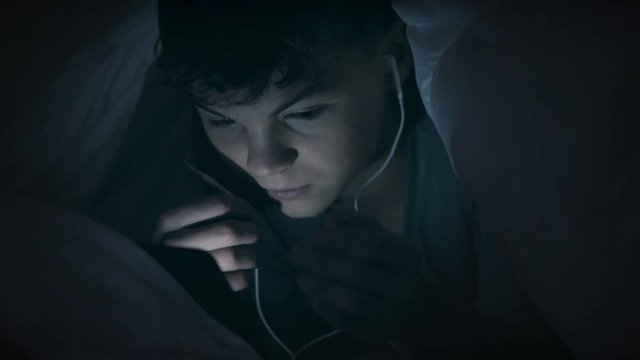 Boy watching movie or playing games on tablet computer at night. Child with headphones lying down under blanket on bed using smartphone or tablet pc. Boy to make video call to talk to friends. 