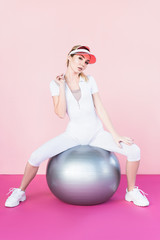 attractive female athlete in visor hat sitting on grey fitness ball and looking at camera on pink