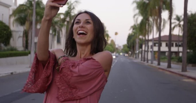Stylish young woman taking smart phone selfies in middle of street with palm trees