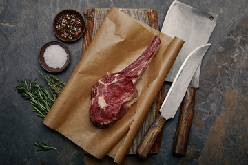 top view of raw rib eye steak on baking paper with kitchen knives, spices, herbs and salt on grey background