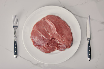 top view of fresh raw meat on plate with kitchen cutlery on white background
