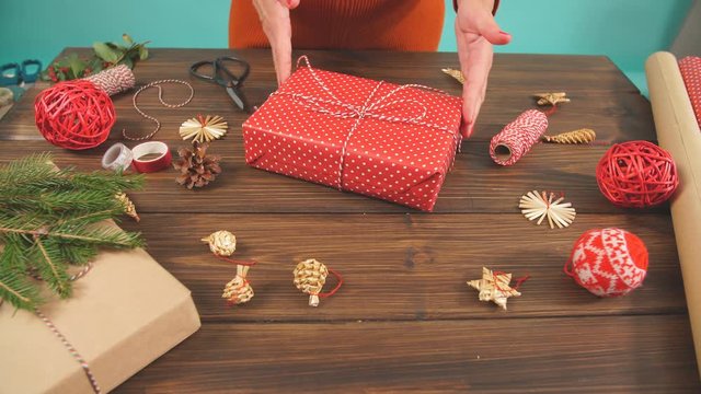 Female hands holding present box wrapped in red paper and tied with backer s twine on the wooden table with decorating elements paper roll, Christmas baubles.