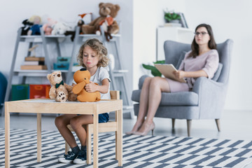 A sad child with trauma playing with toys by a table and a professional psychologist sitting in an...
