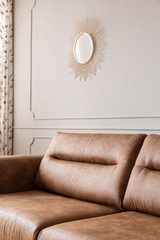 Round gold mirror hanging on the wall with molding in bright sitting room interior with leather brown couch in the real photo