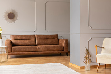 Real photo of open space flat interior with leather brown couch, molding on walls, white carpet and gold decor