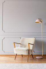 Real photo of pastel pink metal lamp standing by the white armchair in bright sitting room interior with wooden floor, carpet and empty place for your poster on wall with wainscoting