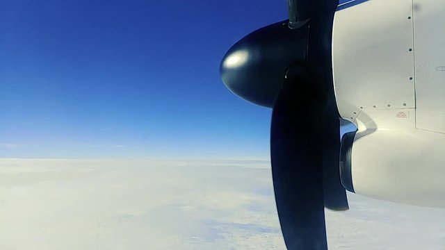 turboprop plane flies in a blue sky over a bed of clouds. From the window of the plane a view of the motor puling the plane forward.
