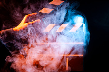 Black and white clapperboard for cinema close up among multicolored red and blue smoke in a man’s hand giving a command to start shooting on a black isolated background