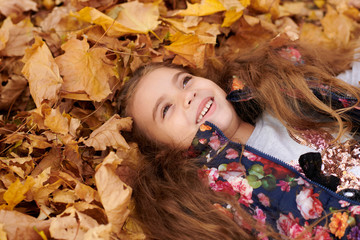 Child girl is lying and playing in fallen leaves in autumn city park.