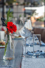 Transparent glass vase with colourful bouquet of flowers against background of table setting in restaurant. Wine and water glasses stand in a row on wooden table. Concept banquet, birthday, conference