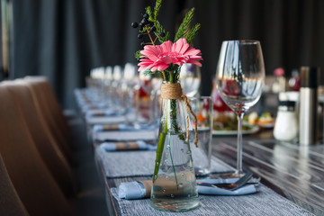 Transparent glass vase with colourful bouquet of flowers against background of table setting in restaurant. Wine and water glasses stand in a row on wooden table. Concept banquet, birthday, conference