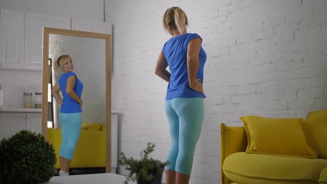 Lovely woman in casual clothes checking her body in mirror, feeling excited and beautiful after successful weight loss program in domestic room. Joyful female admiring her reflection in mirror.