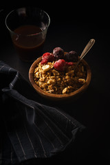 Granola with raspberries and blackberries in wooden bowl with a cup of coffee on black table.