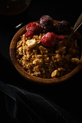 Granola with raspberries and blackberries in wooden bowl with a cup of coffee on black table.