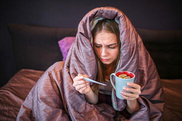 Serious but sick young woman sits on bed with covered blanket. She holds cup of hot tea and...