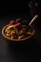 Granola with raspberries and blackberries in wooden bowl with a cup of coffee on black table.