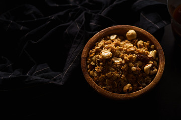 Roasted granola with nuts in wooden bowl with a cup of coffee on black table.