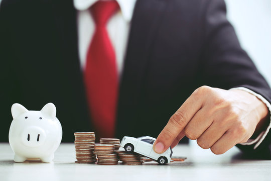 Business Man In Suit Hand Holding Model Of Toy Car White On Over A Lot Money Of Stacked Coins Insurance, Loan And Buying Car Finance Concept. Buy And Installments Down Payment A Car. Piggy Bank Saving