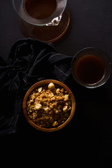 Roasted granola with nuts in wooden bowl with a cup of coffee on black table.