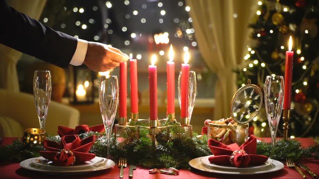 Male hand lights christmas candles. Festival red table setting with garland and Christmas tree in the background Slow motion