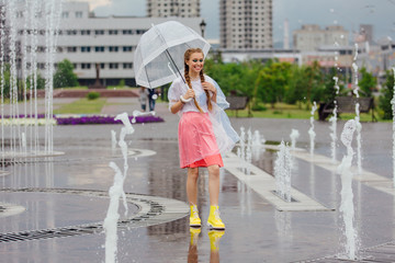 Young pretty girl with two braids in yellow boots and with transparent umbrella stands near fountain.