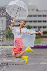 Young pretty girl with two braids in yellow boots and with transparent umbrella jumping near fountain.