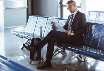 adult businessman sitting at departure lounge at airport and reading newspaper