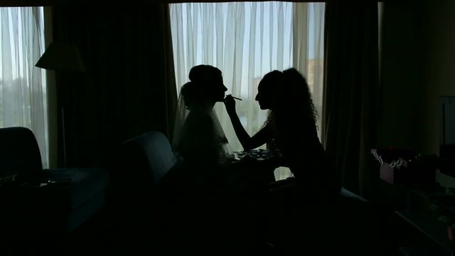Silhouette of bride in the morning in hotel room with makeup artist. Makeup artist making wedding makeup to bride in the morning against the background of a large window, silhouette.