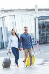 Happy couple of tourists crossing pedestrian, holding hands and pulling their luggage