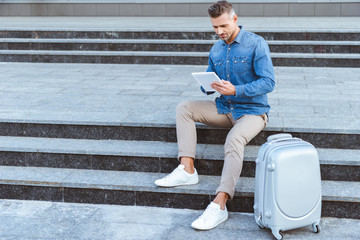 Handsome adult man sitting on the staircase with luggage and using digital tablet