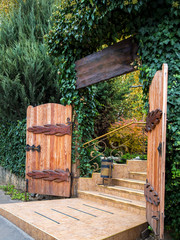 Naklejka premium The entrance to the wooden gate is overgrown with ivy and other evergreens, the passage to the territory of the house is tiled in front of the steps.