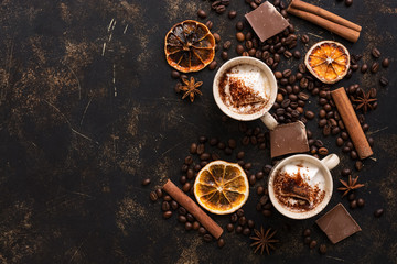 Two cups of hot chocolate on a dark background with roasted coffee beans, cinnamon sticks and sliced orange. Top view, copy space.