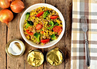 View from above of bowl with tuna risotto with vegetables, tomatoes, broccoli and parsley, onions, cans and brown checkered tablecloth in right frame