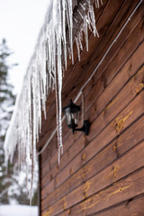 Icicles hang from the roof of a wooden house in the countryside, on a frosty cloudy day.