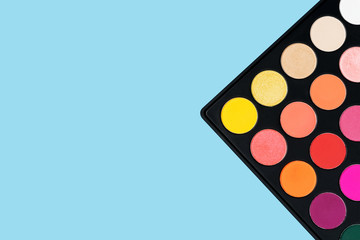Black plastic palette of brightly coloured yellow, red, pink, orange eyeshadow placed in the corner of pastel baby blue background shot with studio light from above