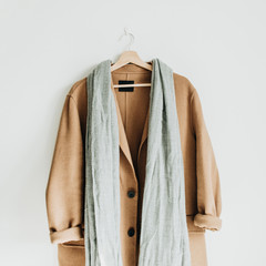 Beige woolen female coat and grey scarf on hanger at white wall. Woman fashion winter cloth concept.